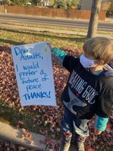 small blonde boy holding sign that says "Dear Adults, I would prefer a future of peace. Thanks!"
