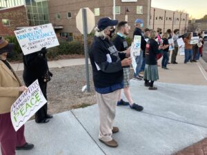 Several protestors with signs that say things about peace standing on sidewalk