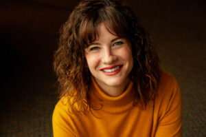 A photo of a woman with medium brown hair and a golden top.