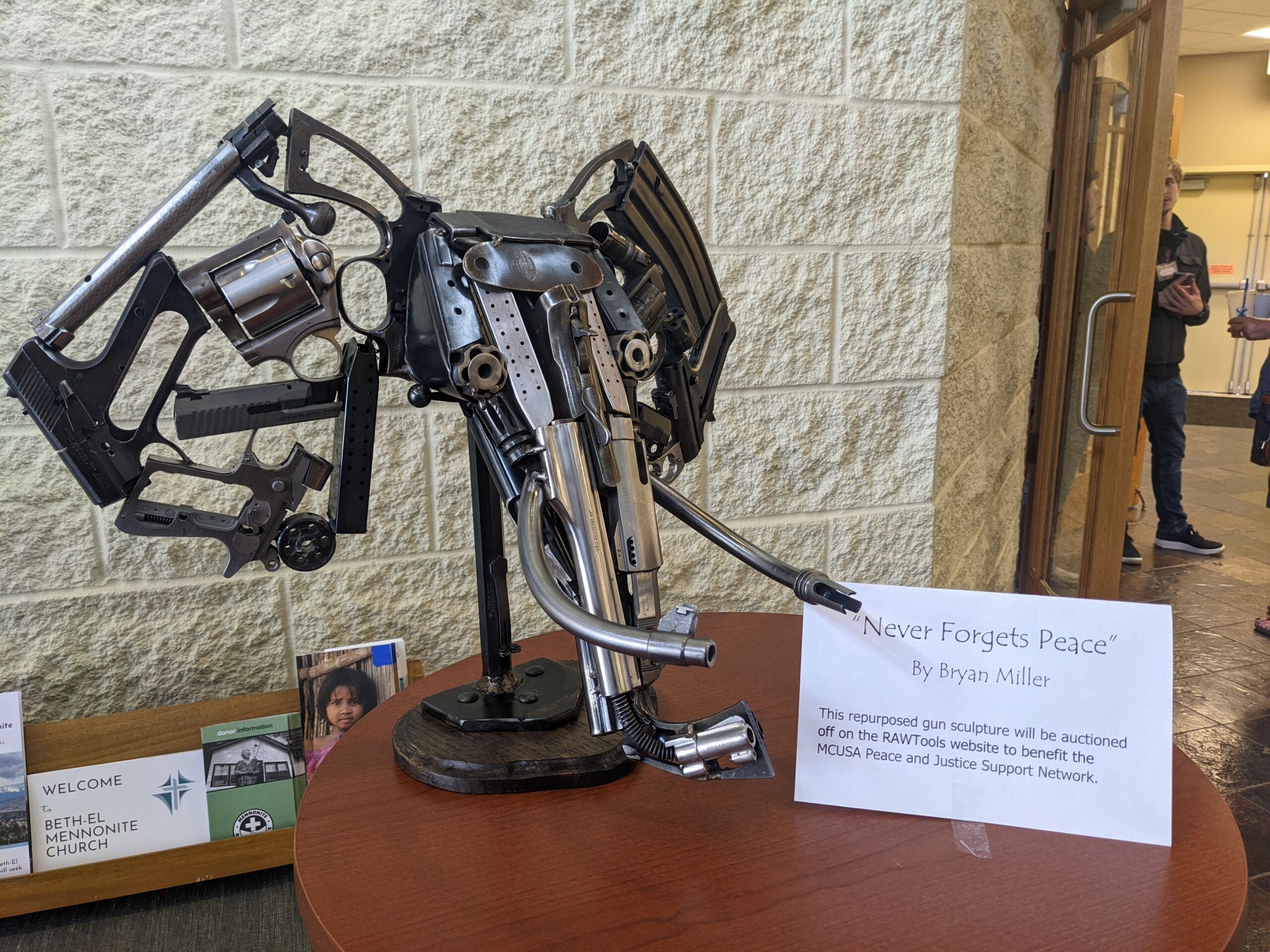 An image of a sculpture made from pieces of disassembled guns. The sculpture resembles an elephant's head and is accompanied by a placard reading, "'Never Forgets Peace' by Brian Miller This repurposed gun sculpture will be auctioned off on the RAWTools website to benefit the MCUSA Peace and Justice Support Network. 