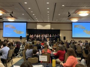 MEC attendees seated at several round tables in a large room, with choral leaders singing on stage at the front of the room.