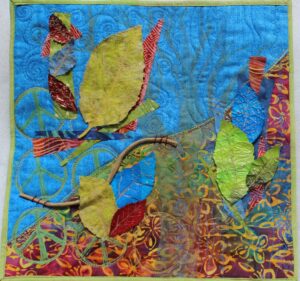 A square quilt with a background split on the diagonal. The top left is blue and the bottom right uses a colorful patterned fabric. In the the foreground are leaves, some made of fabric and others of potato chip bags.
