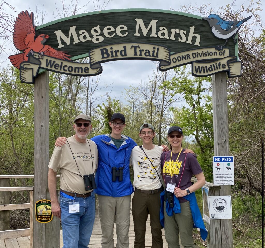 A man, two teenage boys and a woman, all with binoculars, stand under a sign that says "Magee Marsh, welcome Bird Trail, ODNR Division of Wildlife"